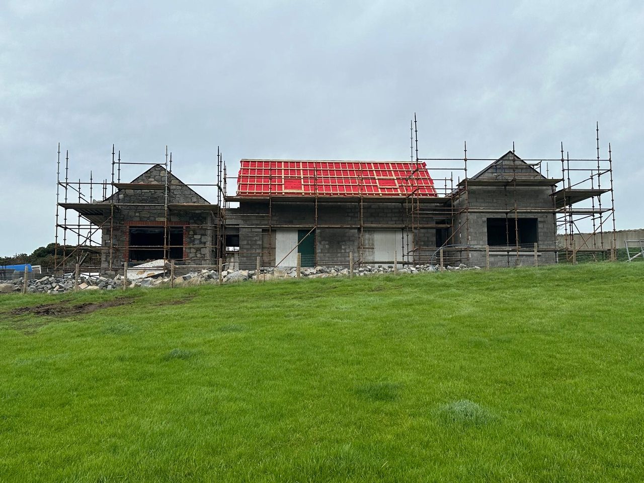 The dwelling under construction behind scaffolding, with stone gables and a new roof, seen across a grass field